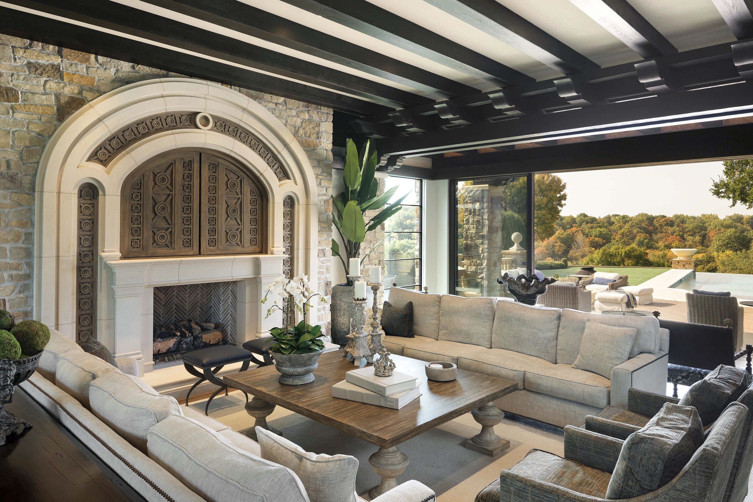 Living room with beautiful wood beam ceilings, stunning fireplace design, and media center with custom hand-carved doors. Stunning view to backyard infinity pool and nature view.