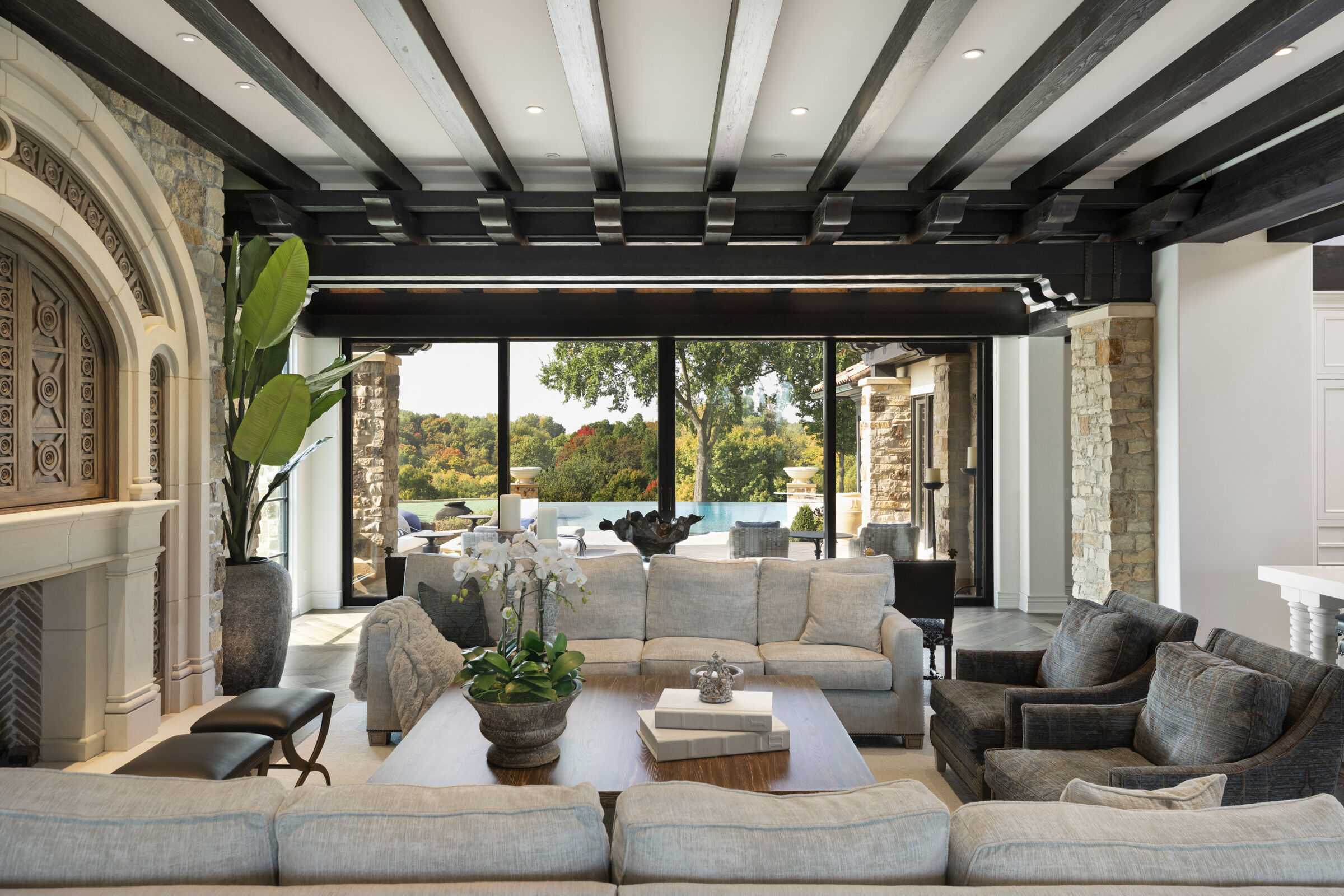 Living room with beautiful wood beam ceilings, stunning fireplace design, and media center with custom hand-carved doors. Stunning view to backyard infinity pool and nature view.