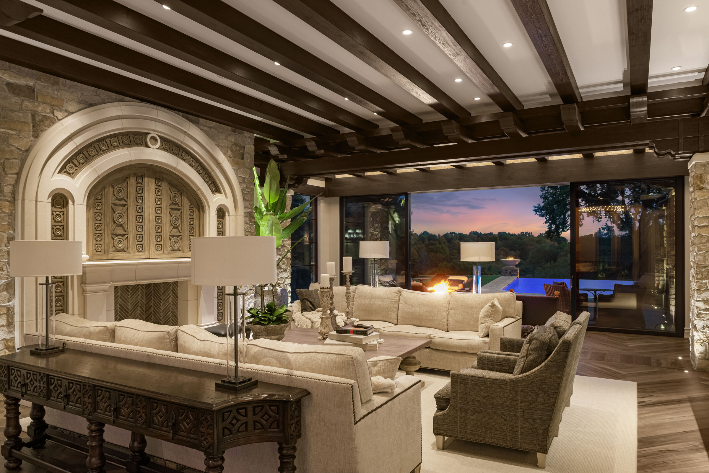 Living room with beautiful wood beam ceilings, stunning fireplace design, and media center with custom hand-carved doors. Stunning view to backyard infinity pool and nature view.