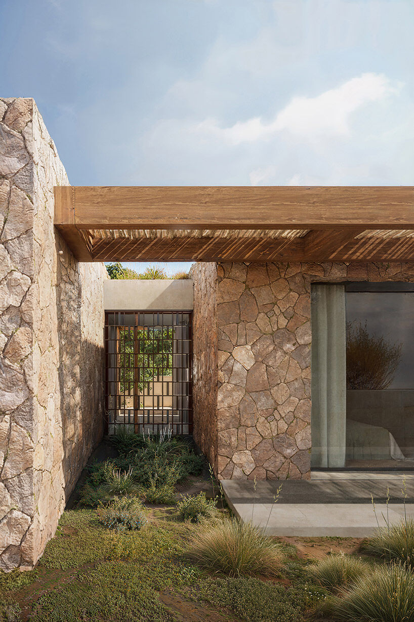 A narrow landscaped passage between stone walls forms a shaded threshold and reinforces the project’s sequence of protected outdoor rooms.
