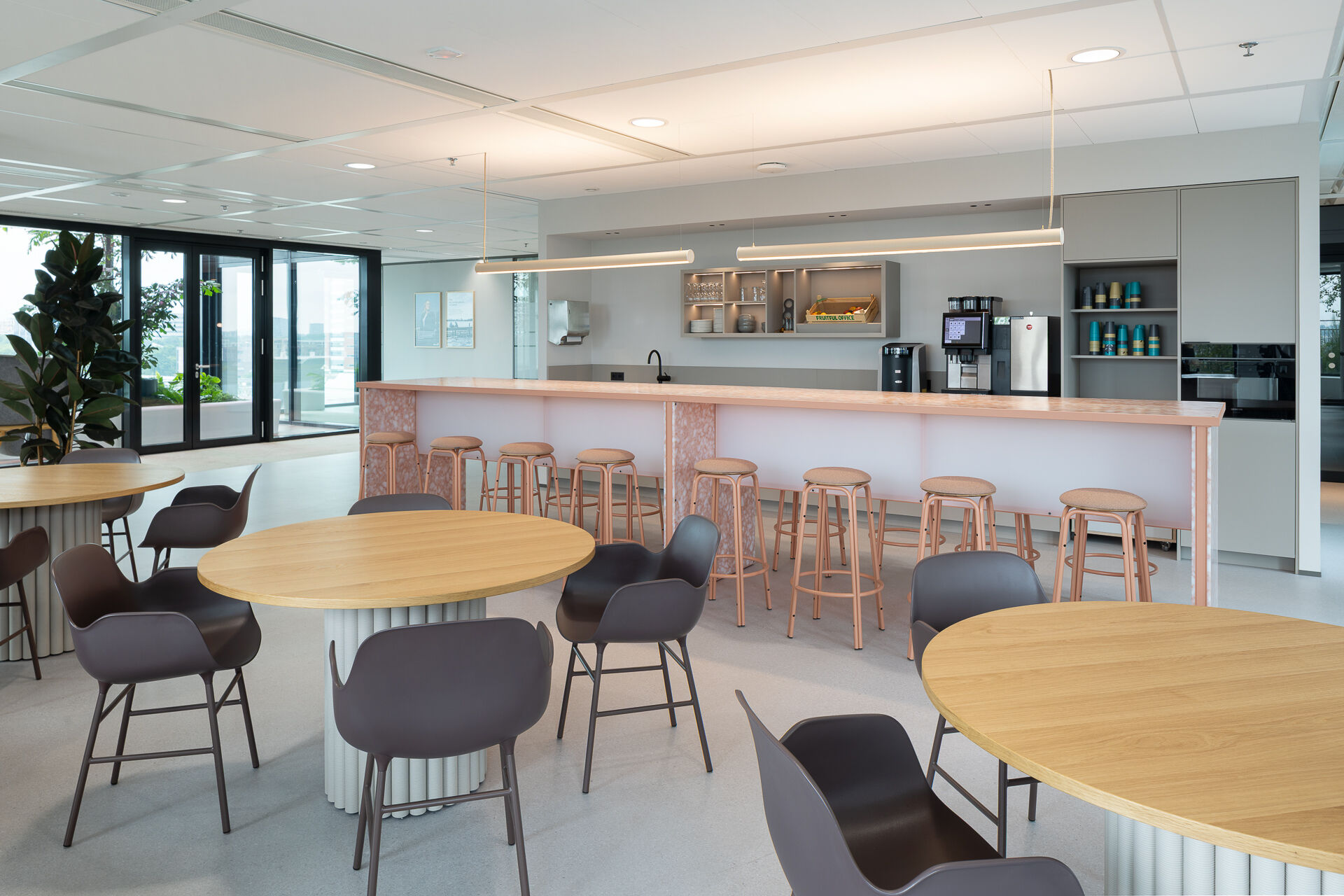 Full overview of the social and café zone, showing the recycled plastic bar counter, kitchen area integrated into the back wall, and the mix of round tables and bar seating. The space functions as both workplace and meeting point.