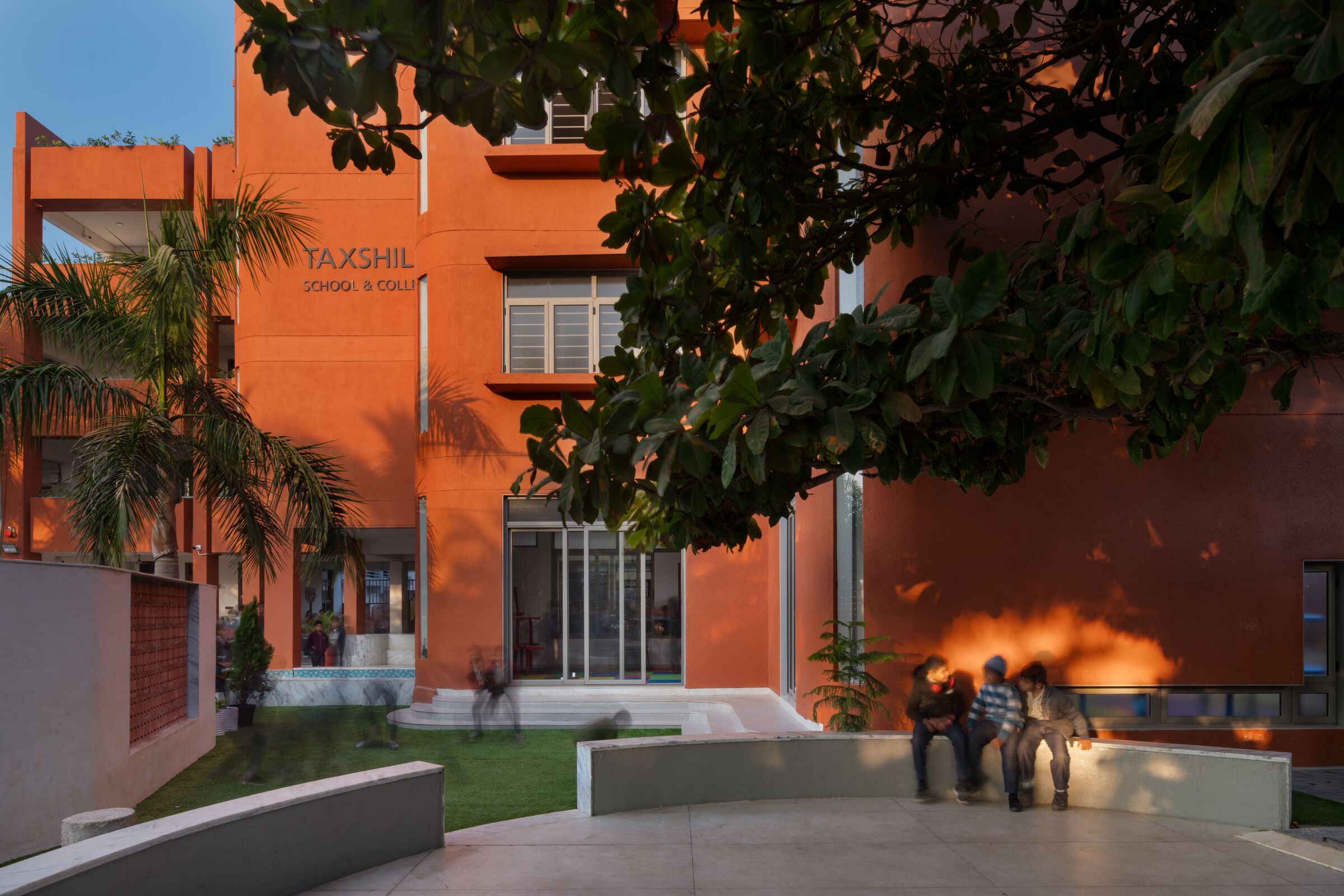 The approach to the new building is shaped as a gradual transition. A tree-lined passage, a courtyard shaded by a badam tree, and a small water body mark the shift from open campus to enclosed architecture