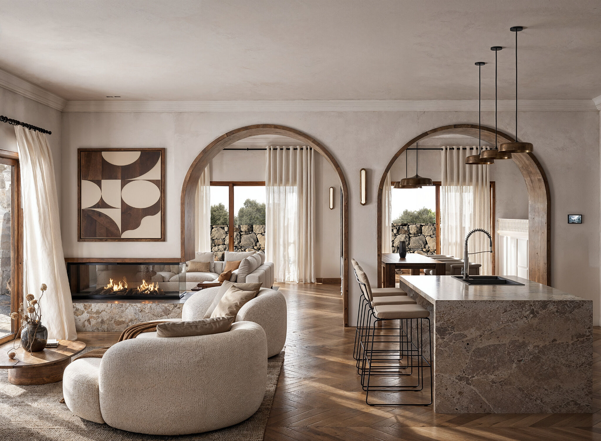 Timber-framed arches connect the kitchen and living areas, while natural stone, warm wood finishes, and a double-sided fireplace establish a cohesive Mediterranean interior.